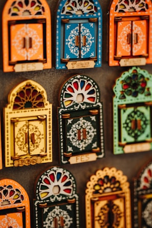 Colorful decorative items resembling miniature window frames are displayed. Each piece is intricately designed with various patterns, featuring bold colors like orange, blue, yellow, and green. Some have small wooden doors with detailed carvings and floral motifs.