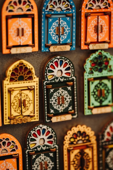 Colorful decorative items resembling miniature window frames are displayed. Each piece is intricately designed with various patterns, featuring bold colors like orange, blue, yellow, and green. Some have small wooden doors with detailed carvings and floral motifs.