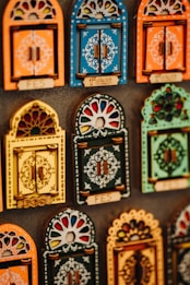 Colorful decorative items resembling miniature window frames are displayed. Each piece is intricately designed with various patterns, featuring bold colors like orange, blue, yellow, and green. Some have small wooden doors with detailed carvings and floral motifs.