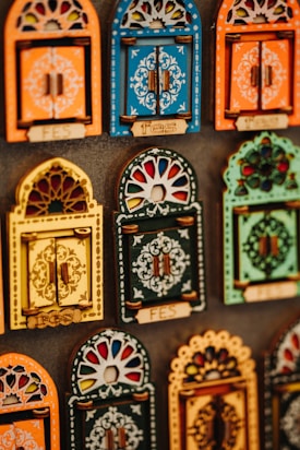 Colorful decorative items resembling miniature window frames are displayed. Each piece is intricately designed with various patterns, featuring bold colors like orange, blue, yellow, and green. Some have small wooden doors with detailed carvings and floral motifs.