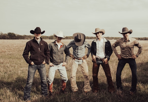 Five individuals wearing cowboy hats and jeans stand in a field with short grass. They each have a relaxed pose and are dressed in western-style clothing. The landscape is open with a horizon line of trees under a cloudy sky.
