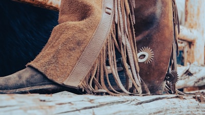 A close-up of a boot with spurs, featuring brown leather chaps with fringe resting against a wooden surface. The scene evokes a rustic, cowboy or Western aesthetic, with the leather textures and wood giving a rugged appearance.