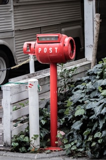A red cylindrical post box is situated next to a white wooden barrier, surrounded by dense green foliage. The box is elevated on a pole and has the word 'POST' written in white capital letters. In the background, there is a vehicle with corrugated siding visible.