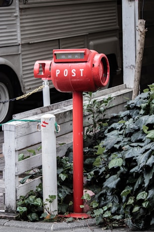 A red cylindrical post box is situated next to a white wooden barrier, surrounded by dense green foliage. The box is elevated on a pole and has the word 'POST' written in white capital letters. In the background, there is a vehicle with corrugated siding visible.