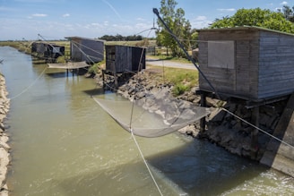 Several wooden fishing huts are situated alongside a calm canal, each equipped with large nets suspended over the water. The huts are elevated on stilts and connected by ropes that hold the fishing nets. The scenery includes grassy areas, trees, and a clear blue sky.
