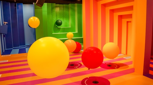 A room with vivid, multicolored walls and floor, featuring large, brightly colored balls in yellow, orange, and red hues. The walls have geometric patterns in pink and orange, while the floor consists of pink and orange stripes. The balls appear to be floating or suspended.