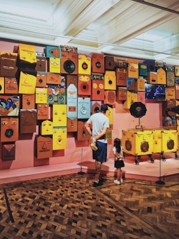 A man and a child stand in front of a vibrant art installation featuring various colorful faces and speaker-like objects mounted on a wall. The faces are painted in different expressions and shades, creating a lively and imaginative display. The room has a parquet floor and is lit by overhead lights, providing a gallery-like ambiance.