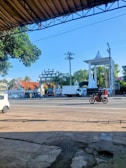 A street scene featuring a busy road with vehicles including a white car, a truck, and a motorcycle carrying two people. In the background, there's a large white monument with a pointed top and a tall pillar. There are trees and a building with an ornate design behind the monument. A blue sky is visible, with overhead power lines crossing the image.