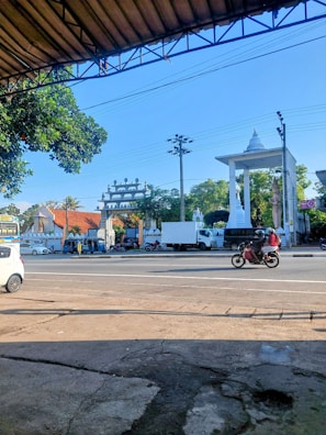 A street scene featuring a busy road with vehicles including a white car, a truck, and a motorcycle carrying two people. In the background, there's a large white monument with a pointed top and a tall pillar. There are trees and a building with an ornate design behind the monument. A blue sky is visible, with overhead power lines crossing the image.