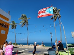 A Puerto Rican flag is prominently displayed against a bright blue sky. Several palm trees sway in the breeze near a coastal area. People are gathered on a sidewalk, with one person sitting at a table near café chairs. Festive string lights are strung across the scene. The ocean is visible in the background, indicating a seaside location.