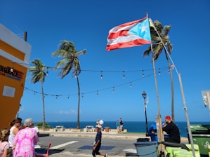 A Puerto Rican flag is prominently displayed against a bright blue sky. Several palm trees sway in the breeze near a coastal area. People are gathered on a sidewalk, with one person sitting at a table near café chairs. Festive string lights are strung across the scene. The ocean is visible in the background, indicating a seaside location.