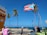 A Puerto Rican flag is prominently displayed against a bright blue sky. Several palm trees sway in the breeze near a coastal area. People are gathered on a sidewalk, with one person sitting at a table near café chairs. Festive string lights are strung across the scene. The ocean is visible in the background, indicating a seaside location.