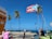 A Puerto Rican flag is prominently displayed against a bright blue sky. Several palm trees sway in the breeze near a coastal area. People are gathered on a sidewalk, with one person sitting at a table near café chairs. Festive string lights are strung across the scene. The ocean is visible in the background, indicating a seaside location.