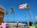 A Puerto Rican flag is prominently displayed against a bright blue sky. Several palm trees sway in the breeze near a coastal area. People are gathered on a sidewalk, with one person sitting at a table near café chairs. Festive string lights are strung across the scene. The ocean is visible in the background, indicating a seaside location.