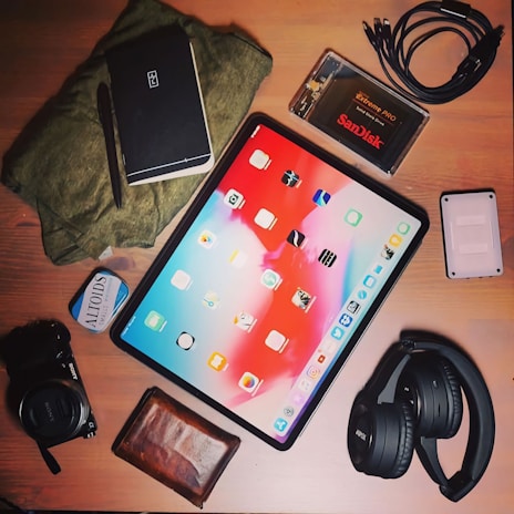 A selection of items lies arranged on a wooden surface. In the center is a tablet displaying a colorful home screen with various app icons. Surrounding it are objects including a black wireless headphone, a digital camera with a lens cap, a small rectangular external hard drive, a SanDisk Extreme Pro SSD in a transparent case, a coiled black cable, a closed black notebook with a pen, a brown leather wallet, a small tin of mints, and a folded green fabric.