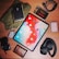 A selection of items lies arranged on a wooden surface. In the center is a tablet displaying a colorful home screen with various app icons. Surrounding it are objects including a black wireless headphone, a digital camera with a lens cap, a small rectangular external hard drive, a SanDisk Extreme Pro SSD in a transparent case, a coiled black cable, a closed black notebook with a pen, a brown leather wallet, a small tin of mints, and a folded green fabric.