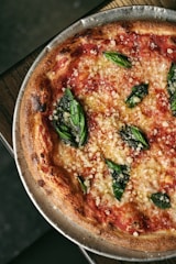 A close-up view of a freshly baked margherita pizza with a crispy, golden-brown crust, topped with rich red tomato sauce, melted cheese, and vibrant green basil leaves. The pizza is served on a metal tray placed on a wooden surface.
