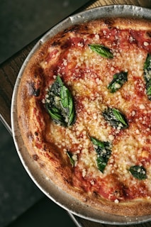 A close-up view of a freshly baked margherita pizza with a crispy, golden-brown crust, topped with rich red tomato sauce, melted cheese, and vibrant green basil leaves. The pizza is served on a metal tray placed on a wooden surface.