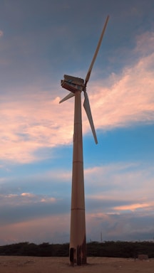 A tall wind turbine stands against a picturesque sky filled with soft pink and blue clouds. The scene is serene and expansive, with the turbine's blades gently angled against the sky. The landscape is flat and dotted with low vegetation, enhancing the focus on the turbine.