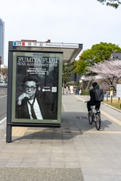 A bus stop with a large advertisement featuring a person in a suit, located near a sidewalk. A cyclist rides past on the right, with a background of trees in full bloom, likely cherry blossoms. Buildings are visible in the vicinity, indicating an urban setting.