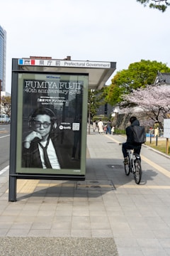 A bus stop with a large advertisement featuring a person in a suit, located near a sidewalk. A cyclist rides past on the right, with a background of trees in full bloom, likely cherry blossoms. Buildings are visible in the vicinity, indicating an urban setting.