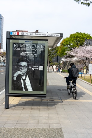A bus stop with a large advertisement featuring a person in a suit, located near a sidewalk. A cyclist rides past on the right, with a background of trees in full bloom, likely cherry blossoms. Buildings are visible in the vicinity, indicating an urban setting.