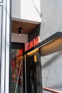 A small entrance to a building features a neon red 'SAUNA' sign above the door. The wall is textured and painted gray, with a smooth white section on top, and there is a black, ceiling-mounted light fixture. The door itself is glass with red circular designs and reflections visible.