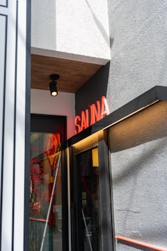 A small entrance to a building features a neon red 'SAUNA' sign above the door. The wall is textured and painted gray, with a smooth white section on top, and there is a black, ceiling-mounted light fixture. The door itself is glass with red circular designs and reflections visible.