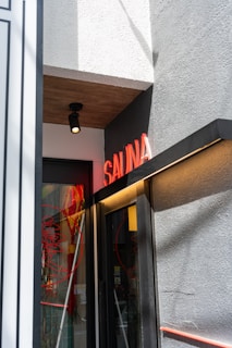 A small entrance to a building features a neon red 'SAUNA' sign above the door. The wall is textured and painted gray, with a smooth white section on top, and there is a black, ceiling-mounted light fixture. The door itself is glass with red circular designs and reflections visible.