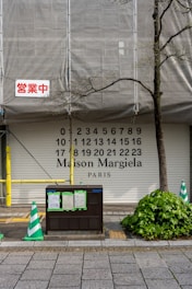 A partially covered storefront with the words 'Maison Margiela PARIS' along with numbers from 0 to 23. There is construction scaffolding with a tarp, a red sign in Japanese, green and white traffic cones, and a bush next to the sidewalk.