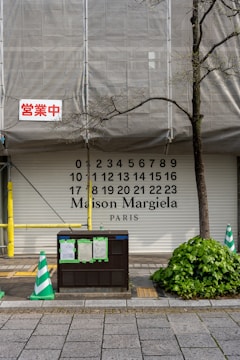 A partially covered storefront with the words 'Maison Margiela PARIS' along with numbers from 0 to 23. There is construction scaffolding with a tarp, a red sign in Japanese, green and white traffic cones, and a bush next to the sidewalk.