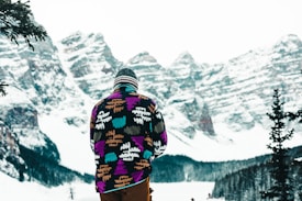 A person wearing a colorful winter jacket and a beanie stands facing a snow-covered mountainous landscape. The jacket features an abstract pattern with vibrant colors such as purple, teal, and orange. In the background, there are towering mountains capped with snow and coniferous trees on the lower slopes.