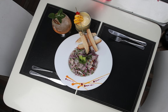 A plate of salad with finely chopped vegetables is garnished with dressing and accompanied by breadsticks. Beside it, there are two drinks; one is a light-colored smoothie topped with slices of fruit and a cherry, the other is a cocktail with mint leaves. The table is set with two forks and two knives on black placemats.