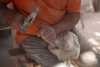 A person is carving a piece of wood or stone with a chisel and a hammer. The focus is on their hands and the partly sculpted object they are working on, while wearing a dusty orange shirt and pants suggesting manual labor.