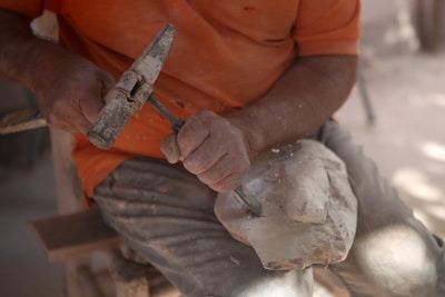 A person is carving a piece of wood or stone with a chisel and a hammer. The focus is on their hands and the partly sculpted object they are working on, while wearing a dusty orange shirt and pants suggesting manual labor.