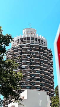 A high-rise building with a unique, geometric design dominates the scene. The structure features multiple angular balconies that create a zigzag pattern. At the top, there is a circular section with protruding elements, giving it a crown-like appearance. Surrounding the building is a clear blue sky, with trees partially visible in the foreground, adding a natural element to the urban landscape.