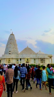 A large group of people is gathered around an intricately designed white marble temple with ornate domes and spires. The temple exterior features detailed carvings and decorative patterns. The sky above is partly cloudy with a hint of evening light.