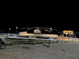 A helicopter is displayed on a platform surrounded by lights arranged in a semicircle pattern. The area is dimly lit, with lamp posts providing some light. There is a pedestrian walking nearby and construction materials scattered around. The background is dark, emphasizing the lit platform and helicopter.