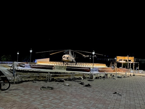 A helicopter is displayed on a platform surrounded by lights arranged in a semicircle pattern. The area is dimly lit, with lamp posts providing some light. There is a pedestrian walking nearby and construction materials scattered around. The background is dark, emphasizing the lit platform and helicopter.