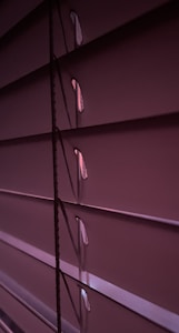 Horizontal window blinds are shown in a dimly lit setting with subtle gradients of purple and pink tones. Light peeks through the small openings between the slats.