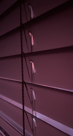 Horizontal window blinds are shown in a dimly lit setting with subtle gradients of purple and pink tones. Light peeks through the small openings between the slats.