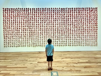 A child stands in a spacious gallery in front of a large wall installation made up of rows of small, red-tinted sculptural forms. The floor is polished wood, and the room is well-lit, creating a serene atmosphere.