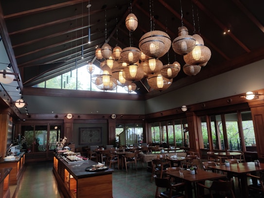 A dining area with a high ceiling and a large number of wicker pendant lights creating a warm, inviting atmosphere. Wooden tables and chairs are neatly arranged in the room, and large windows allow natural light to filter in, offering a view of greenery outside. A buffet setup is visible on the left side with various dishes.