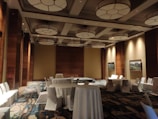 A spacious, well-lit conference room with high ceilings adorned with circular light fixtures. The room is decorated with a mix of warm wooden panels and neutral-colored walls. Several round tables covered with white tablecloths are surrounded by white chairs. The floor features a patterned carpet, and framed artwork hangs on the walls.
