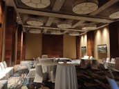 A spacious, well-lit conference room with high ceilings adorned with circular light fixtures. The room is decorated with a mix of warm wooden panels and neutral-colored walls. Several round tables covered with white tablecloths are surrounded by white chairs. The floor features a patterned carpet, and framed artwork hangs on the walls.