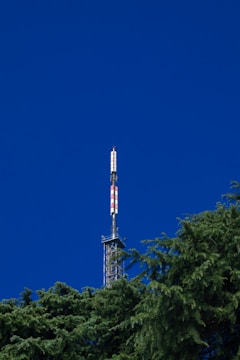 A communication tower with a series of red and white stripes extends into a vibrant blue sky. The base of the tower is surrounded by lush green pine trees.