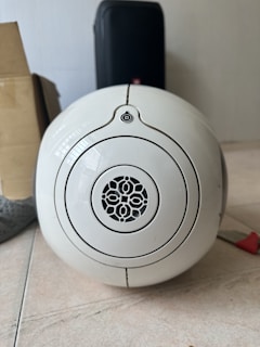 A white, spherical speaker with a decorative grille pattern occupies the center of the frame, resting on a tiled floor. The background includes a cardboard box to the left and a black, rectangular suitcase behind the speaker. Part of a sock and a red-handled object are visible on the floor.
