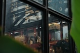 A view through a window into a cafe or kitchen area, with menu items written on a board displaying various drink options such as Tropical and Oriental. Inside, there are shelves with various items including a blender, kettles, cartons, and glassware. Large green leaves are partially visible, framing the edge of the image.