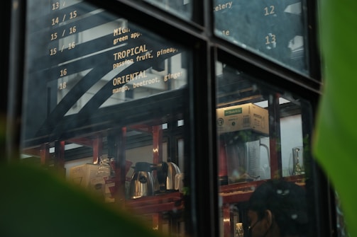 A view through a window into a cafe or kitchen area, with menu items written on a board displaying various drink options such as Tropical and Oriental. Inside, there are shelves with various items including a blender, kettles, cartons, and glassware. Large green leaves are partially visible, framing the edge of the image.