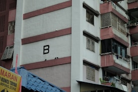 High-rise residential building with pink and white exterior. A large letter 'B' is visible on one of the walls. The building has multiple balconies secured with white grills. The lower portion includes a blue roof and a yellow sign with text in a different language.